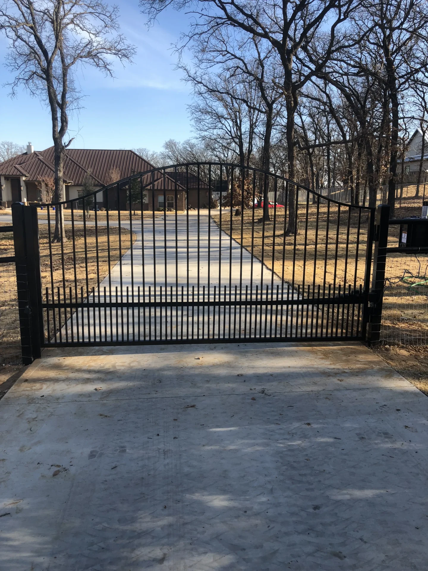 Black wrought iron driveway gate in front of a house, surrounded by trees and a clear blue sky